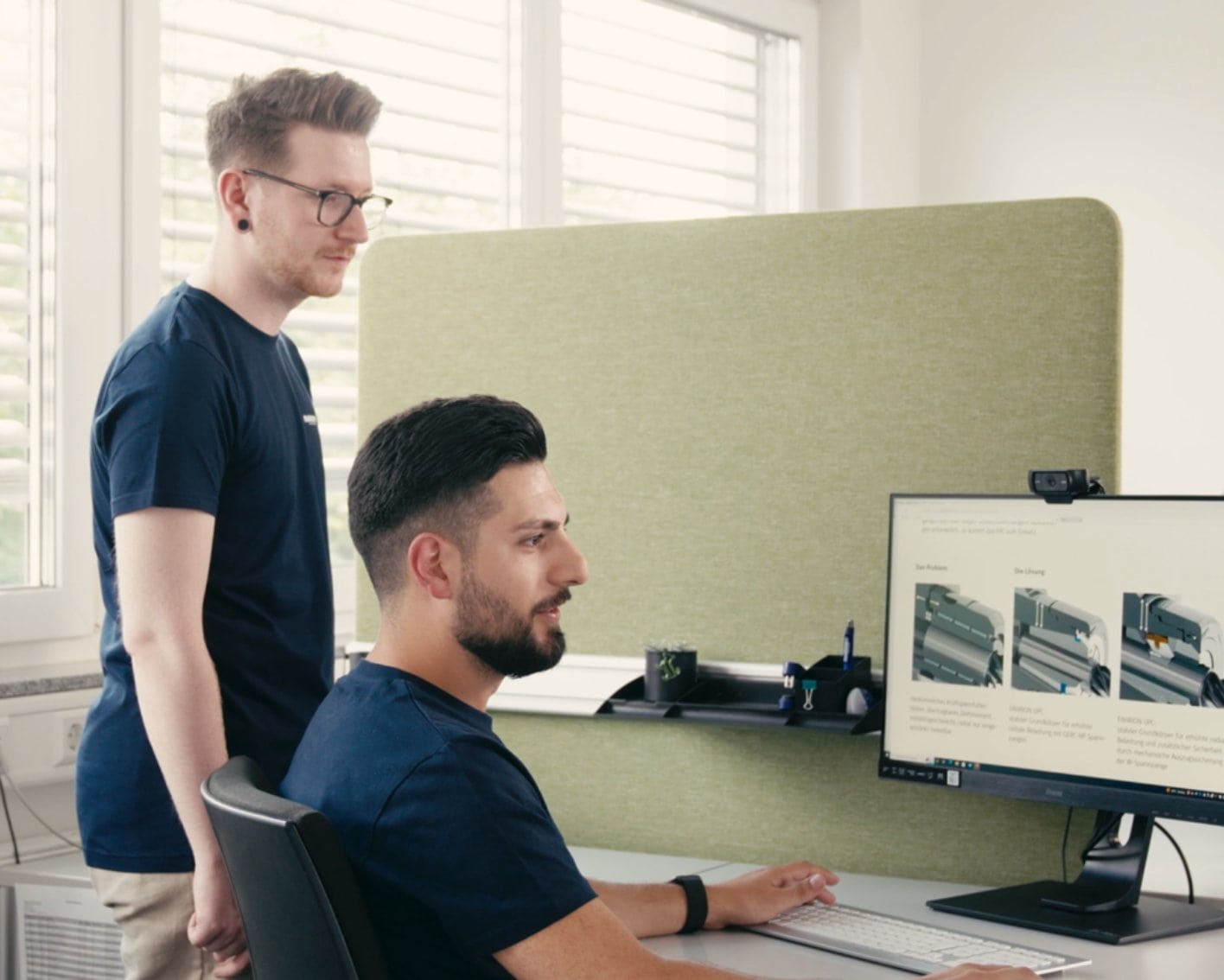 Two men in an office are looking at a computer screen, one sitting at a desk, the other standing next to it. Technical drawings can be seen on the screen.