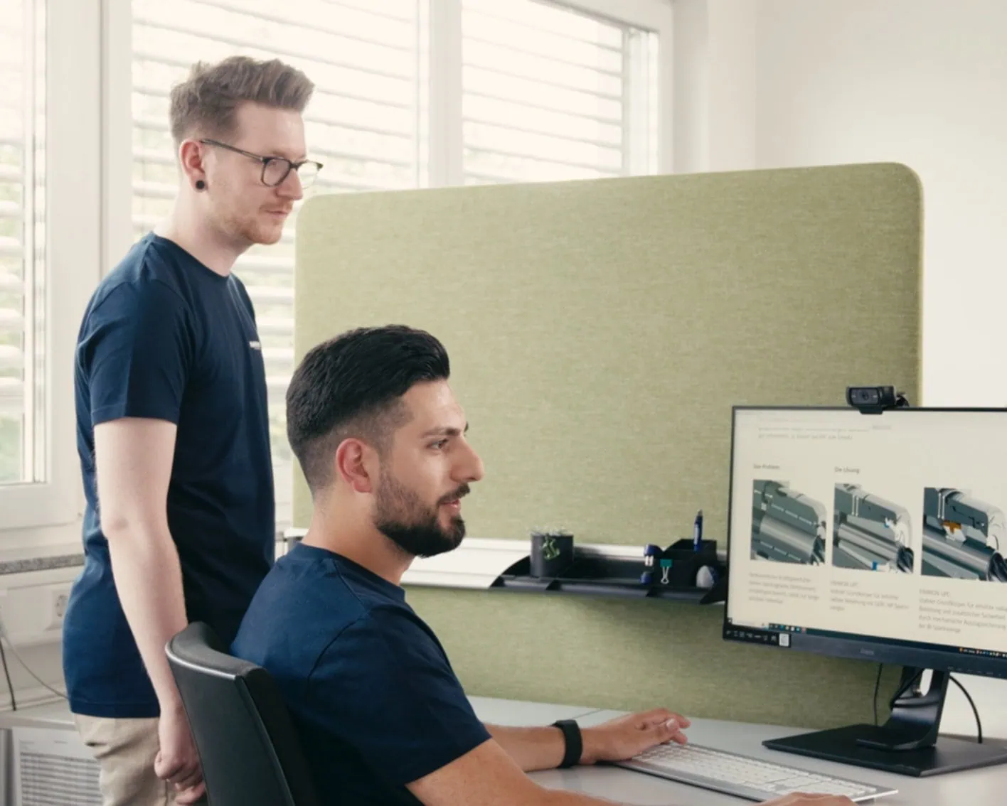 Zwei Männer in einem Büro schauen auf einen Computerbildschirm, einer sitzt am Schreibtisch, der andere steht daneben. Auf dem Bildschirm sind technische Zeichnungen zu sehen.