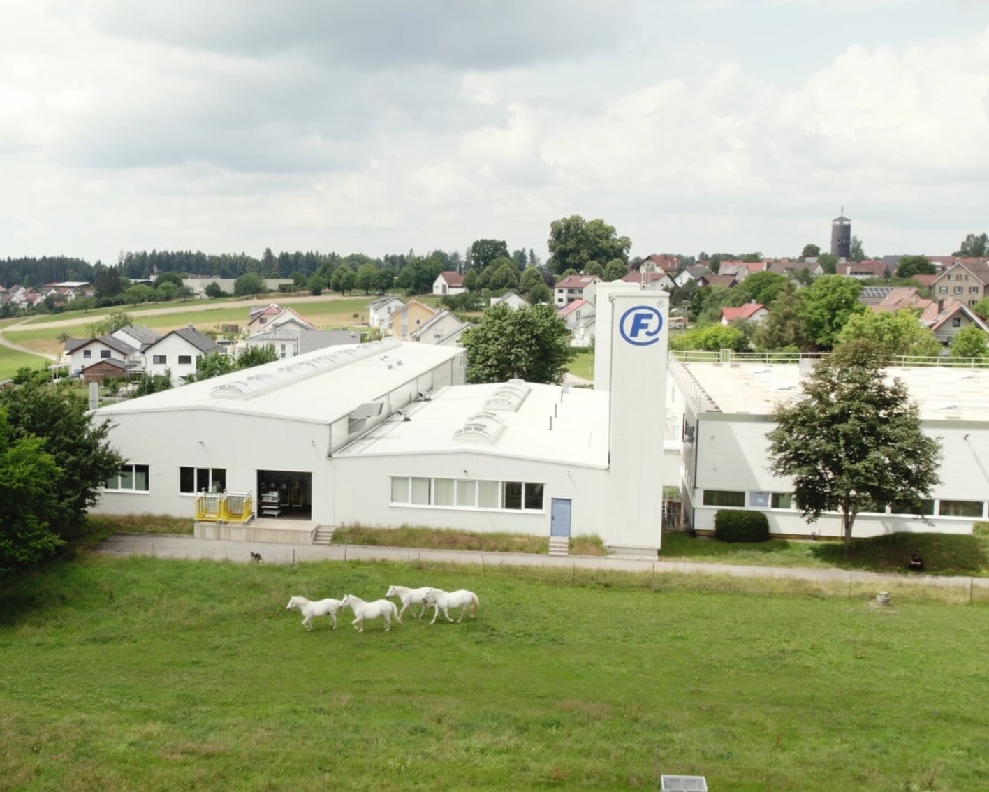 Aerial shot of the white FAHRION industrial facility with the “F” emblem on one section of the façade. Four white horses are running across the meadow in front of the building.