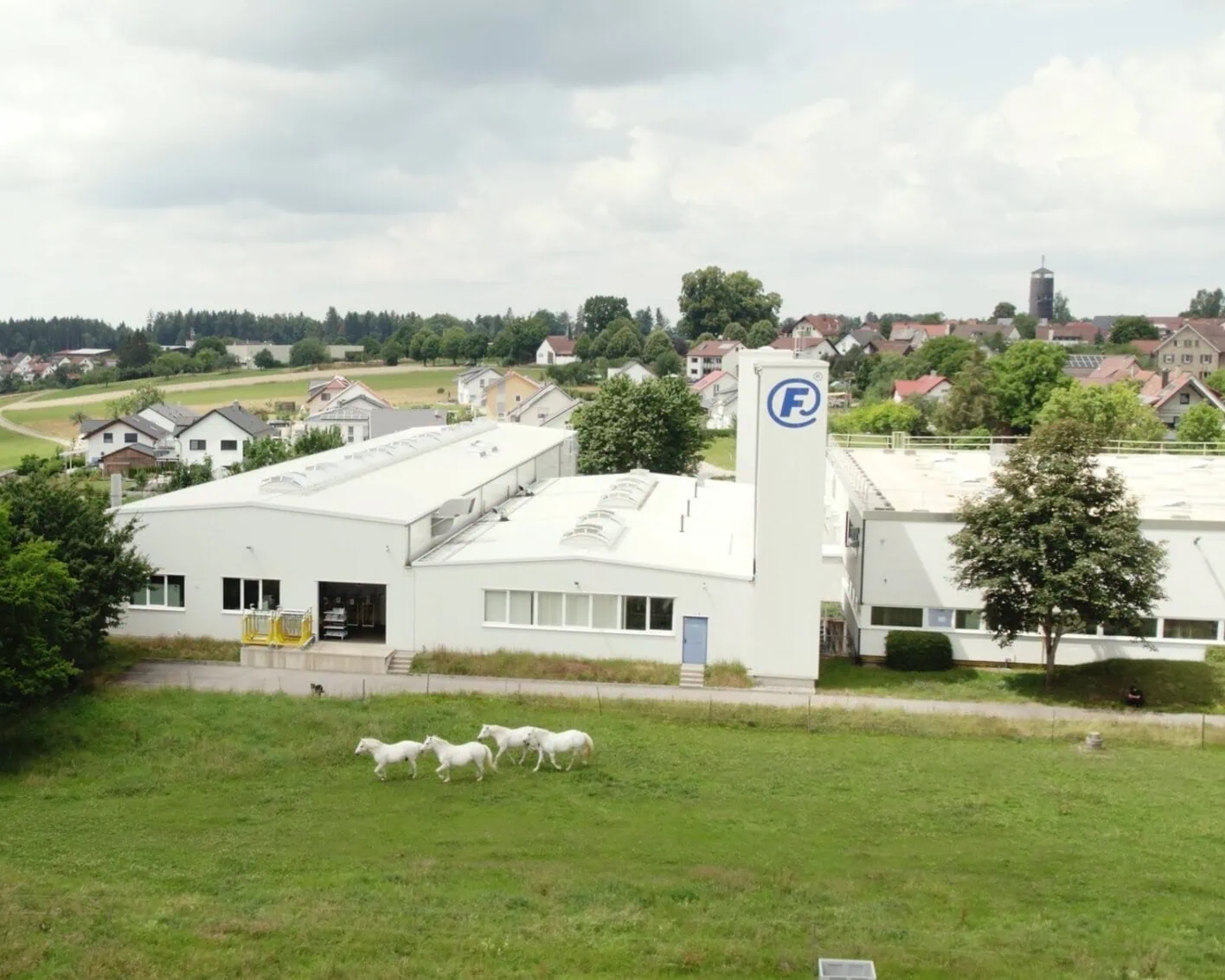 Luftaufnahme des weißen Industriegebäudes von FAHRION mit der Bildmarke 'F' auf einem Gebäudeteil. Vor dem Gebäude laufen vier weiße Pferde auf einer Wiese.