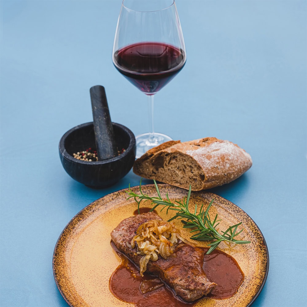A plate with a steak garnished with onions and rosemary, next to it a glass of red wine, a piece of bread, and a mortar with peppercorns.