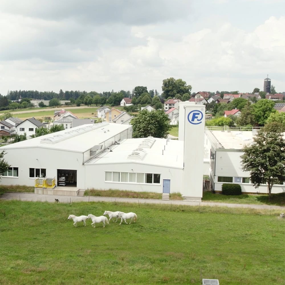 Aerial shot of the white FAHRION industrial facility with the “F” emblem on one section of the façade. Four white horses are running across the meadow in front of the building.