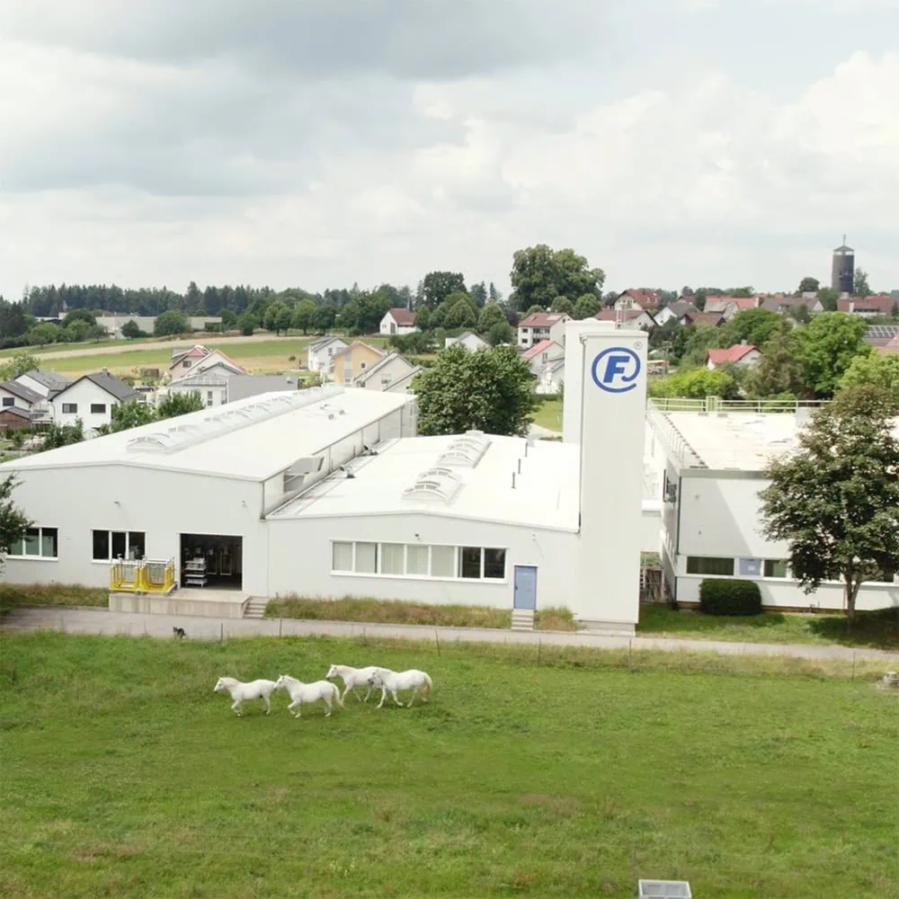 Luftaufnahme des weißen Industriegebäudes von FAHRION mit der Bildmarke 'F' auf einem Gebäudeteil. Vor dem Gebäude laufen vier weiße Pferde auf einer Wiese.